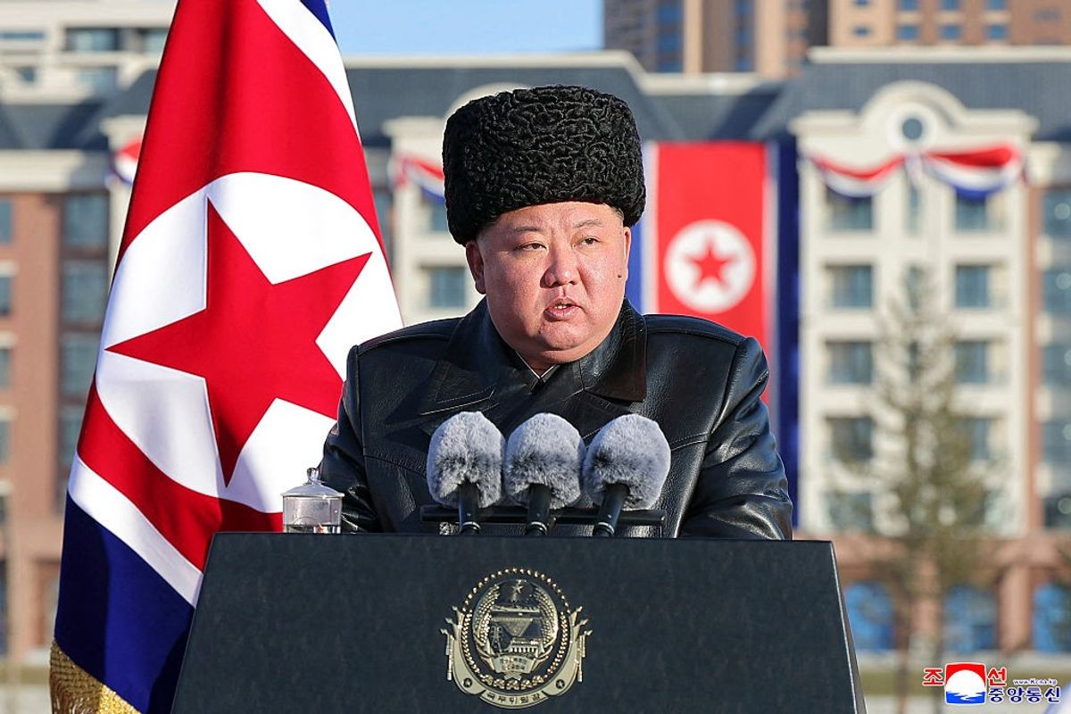 Man in a fur hat speaks at a podium with microphones, North Korean flag in the background.