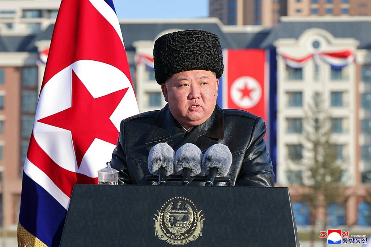 Man in a fur hat speaks at a podium with microphones, North Korean flag in the background.