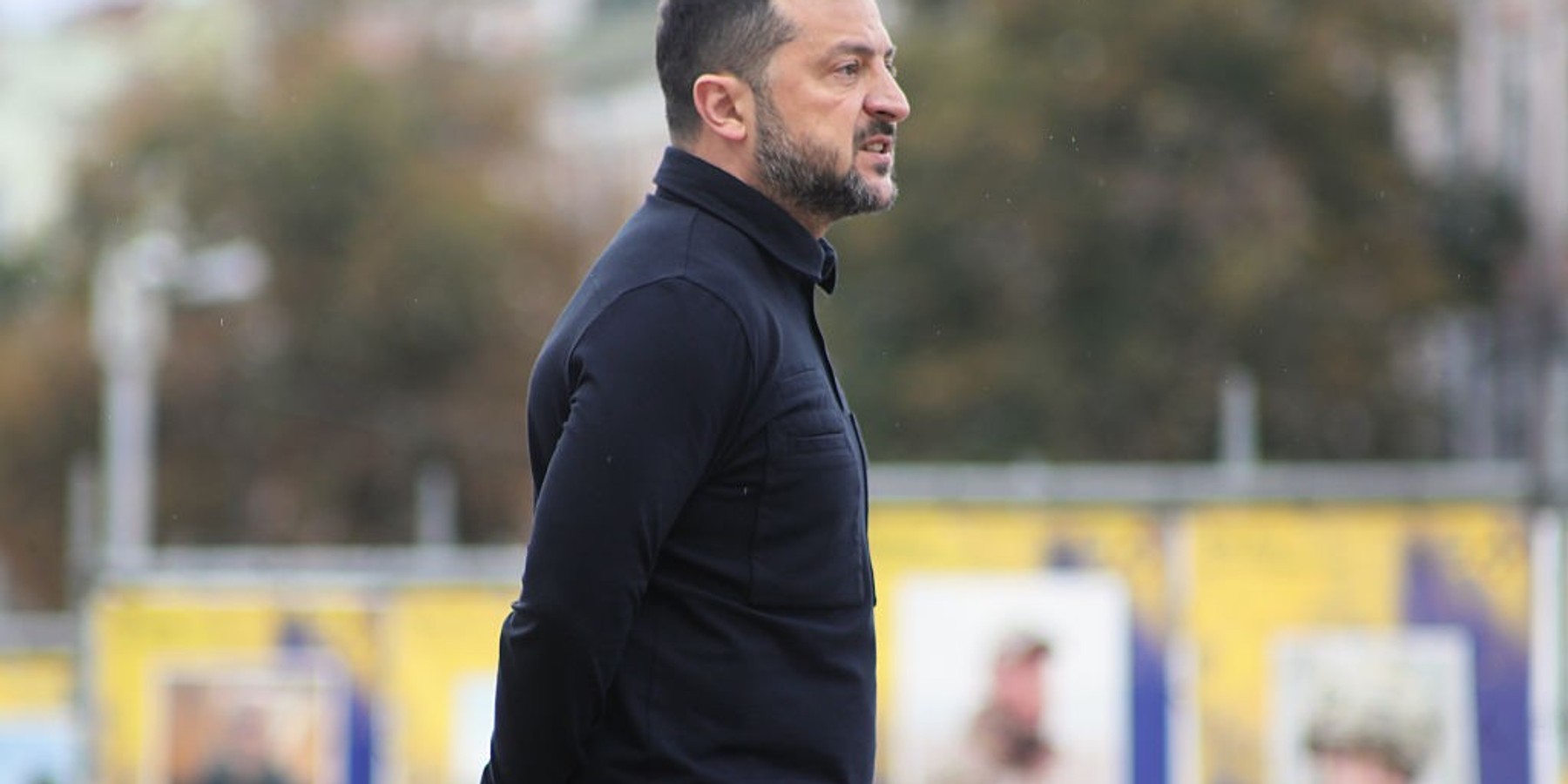 Man in black jacket standing outdoors with blurred background.