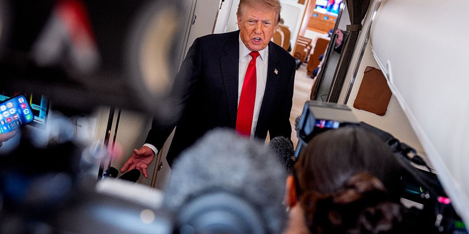 Person speaks to media on an airplane, surrounded by cameras and microphones.