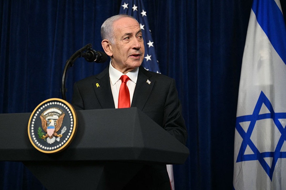 Politician speaks at podium with American and Israeli flags in the background.