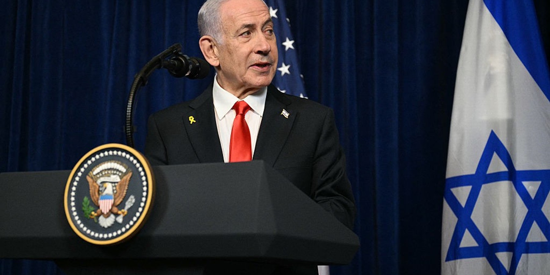 Politician speaks at podium with American and Israeli flags in the background.
