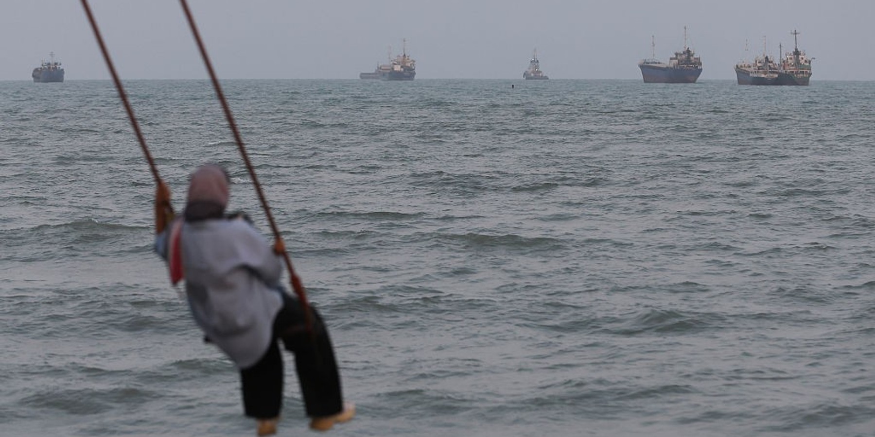Strait Of Hormuz Remains Effectively Closed As Iran Makes New Demands For Reopening Of Waterway BANDAR ABBAS, IRAN - AUGUST 10: A person swings over the shoreline as ships are seen anchored in the Strait of Hormuz on August 10, 2026 off the coast of Bandar Abbas, Iran. In a recent statement, Iran's Supreme National Security Council said the vital waterway would not reopen until the United States corrects its behavior, demanding that the U.S. end its war on Iran and compensate the country for war damage, among other conditions. Iran has largely halted vessel traffic through the strait with the threat of strikes; meanwhile, the U.S. has imposed a blockade on Iranian ports. (Photo by Ali Saeedi/Getty Images)