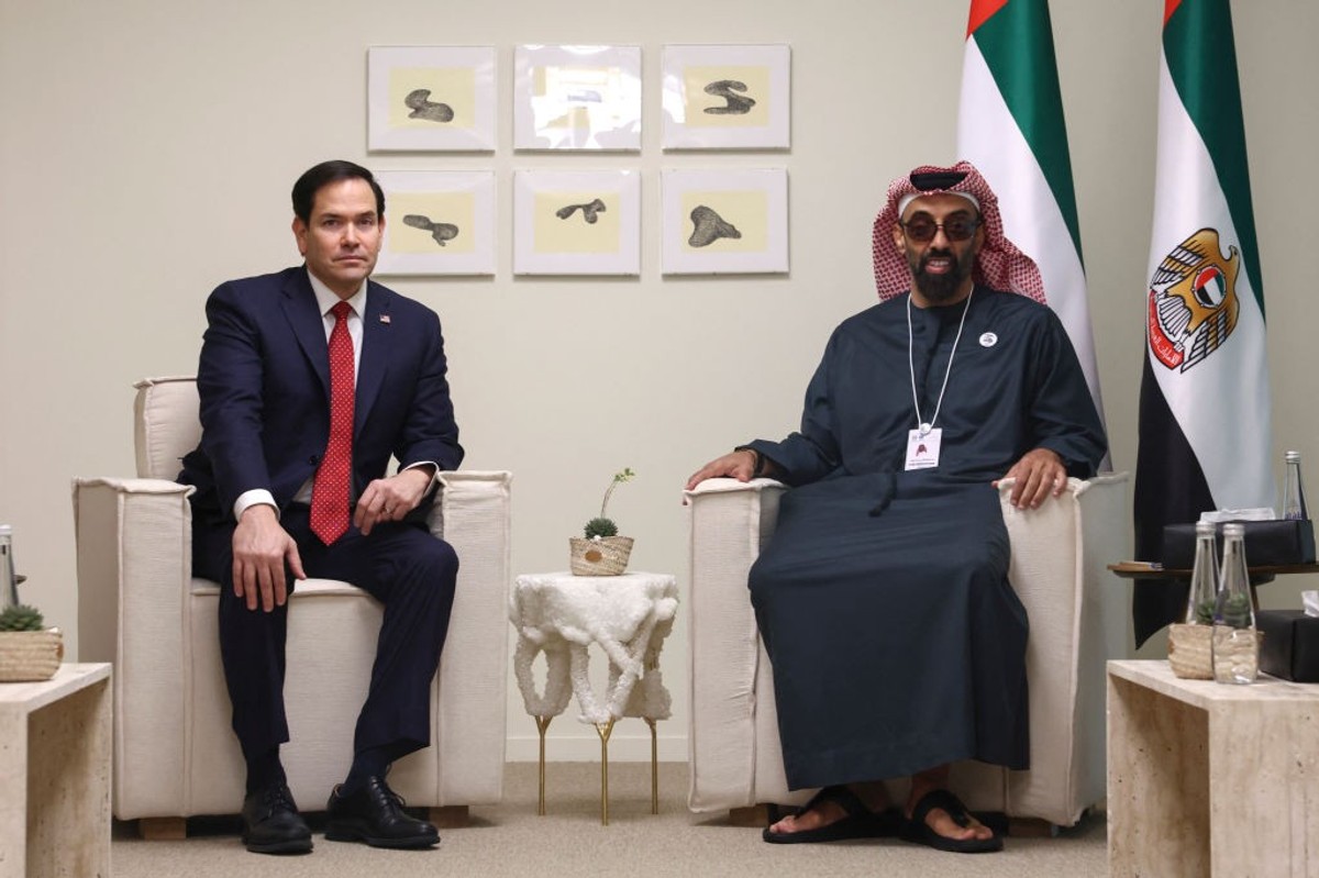 Two men sitting in chairs, with UAE flags and framed art on the wall behind them.