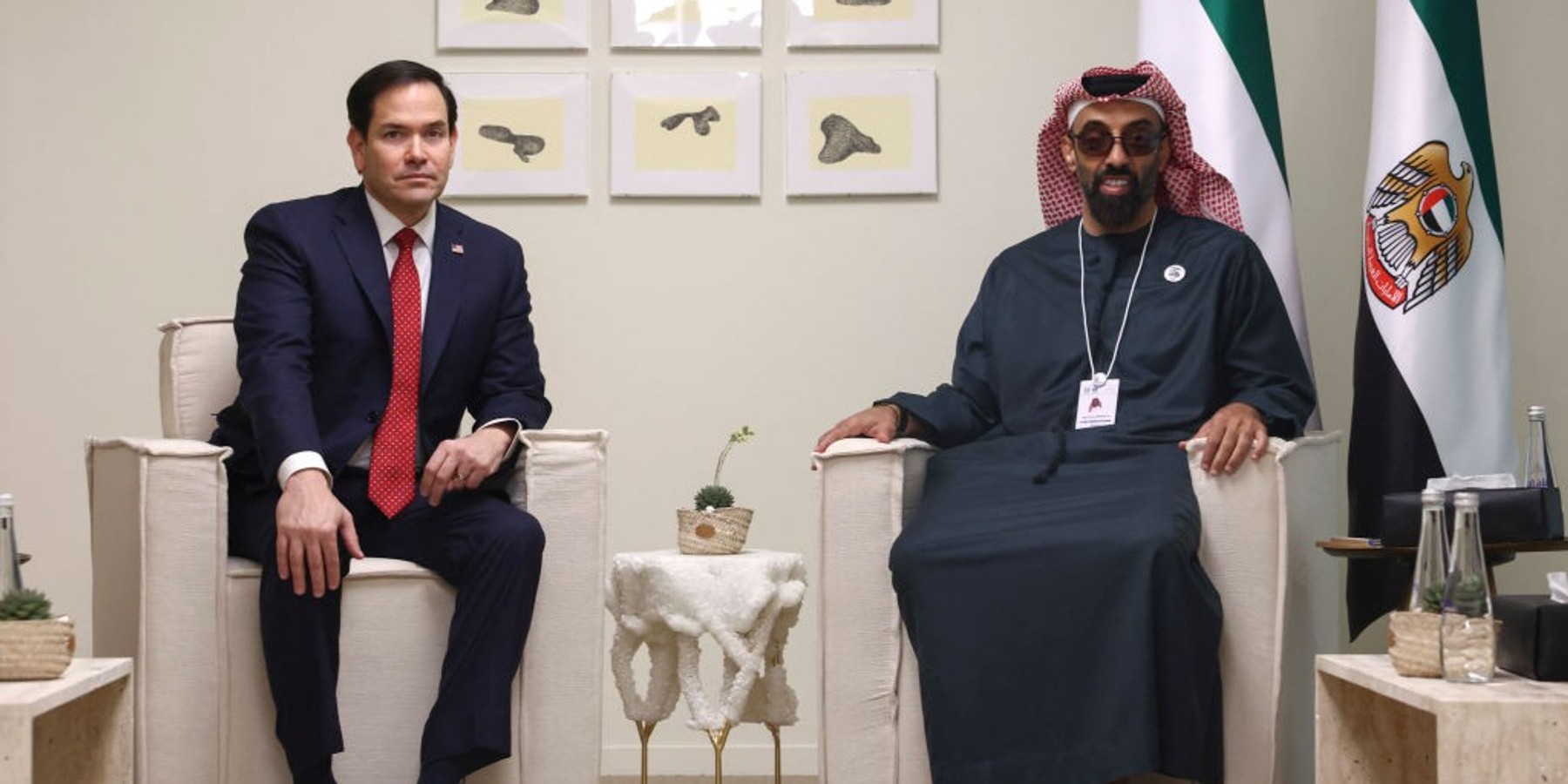 Two men sitting in chairs, with UAE flags and framed art on the wall behind them.
