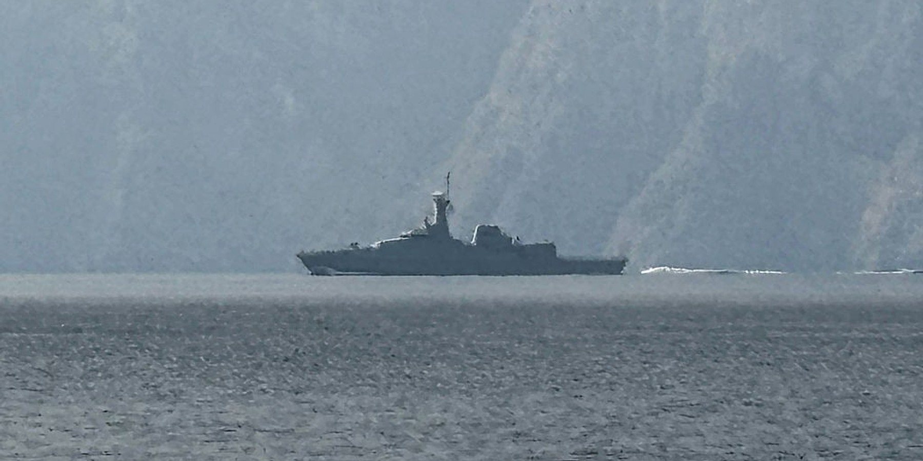 Warship sailing in misty waters with cliffs in the background.