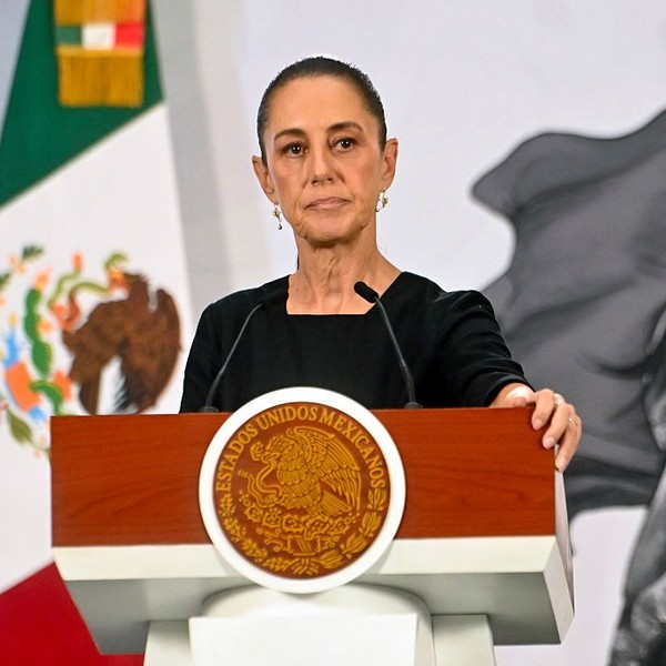 Woman speaking at podium with Mexican seal, flag, and artwork in background.